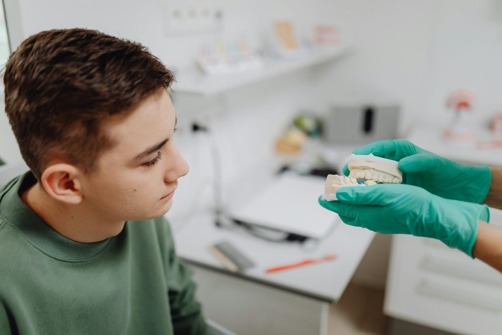 Teenager attending a dental consultation, examining a dental model held by a dentist wearing green gloves.